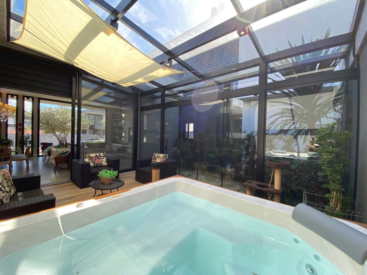 A spacious sunroom is highlighted, featuring a hot tub surrounded by large glass panels that invite natural light. Comfortable seating areas with floral cushions are visible, along with greenery that enhances the tranquil ambiance of the space.