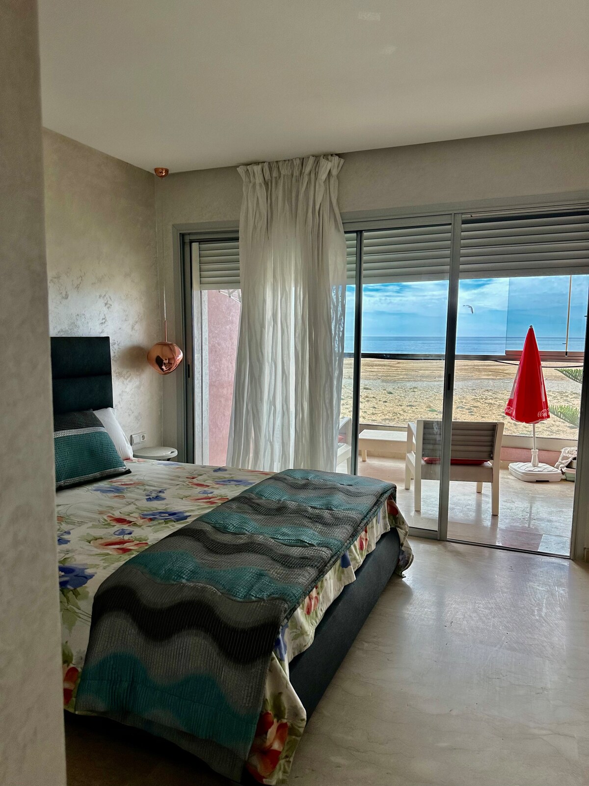 A bright bedroom features a spacious bed with patterned bedding. Light-colored walls enhance the airy feel, while large sliding glass doors open to a seaside view, revealing sandy beach and patio area with an umbrella.