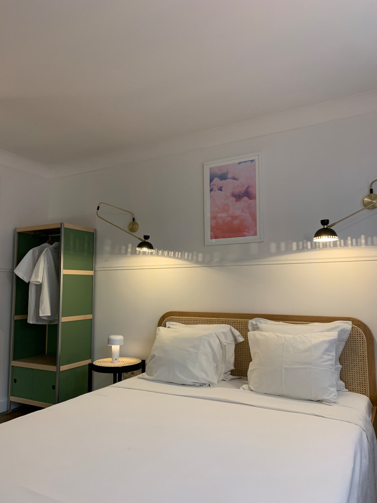 A bright sleeping area features a spacious bed with crisp white linens and two matching pillows. Decorative sconces provide soft lighting flanking the bed. A colorful piece of art hangs above, while a minimalist wardrobe with an adjoining side table is present beside the bed.