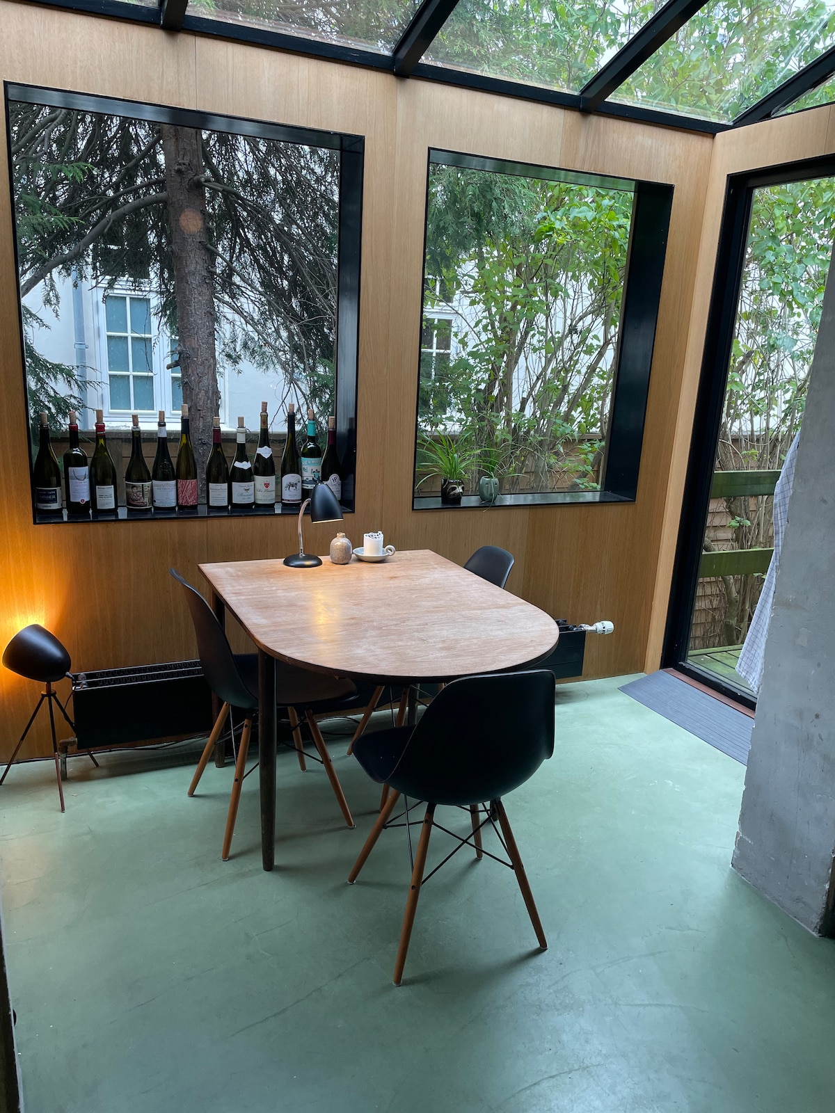 A well-lit dining area features a round wooden table surrounded by four black chairs. Natural light enters through large windows, highlighting the greenery outside. Shelves display an assortment of wine bottles, complementing the space's inviting atmosphere.