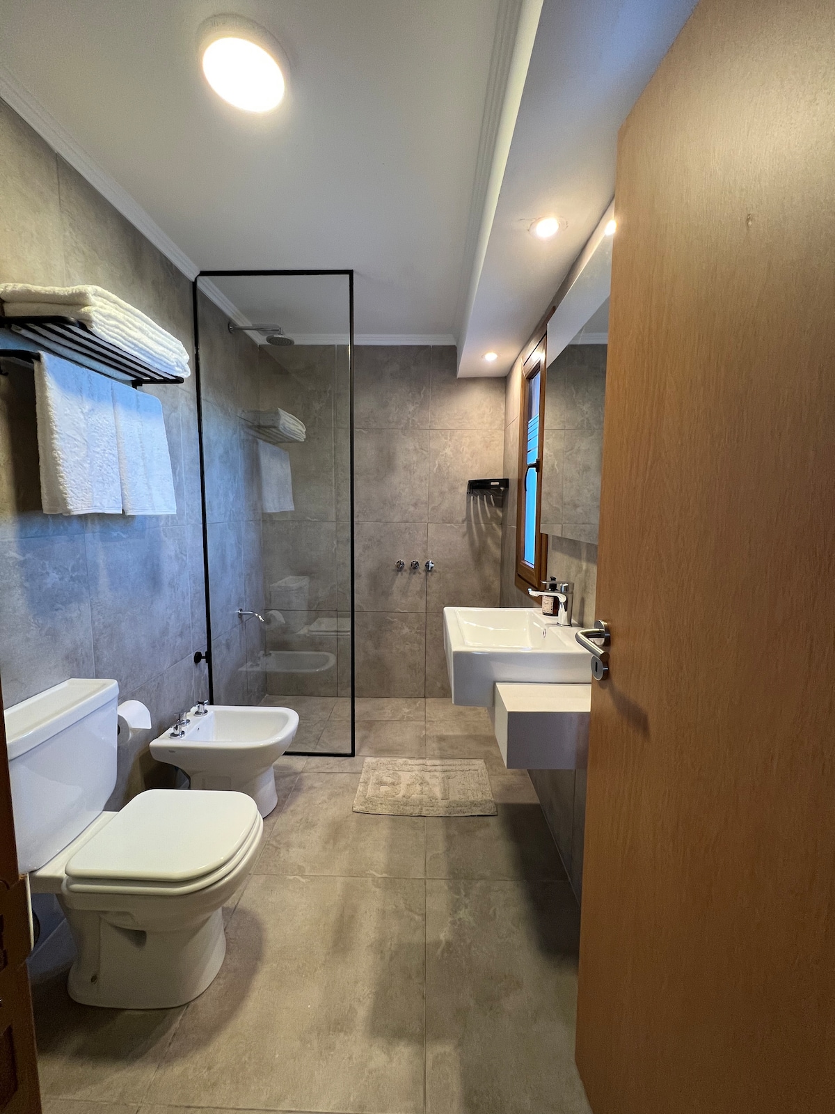 The bathroom features modern fixtures with a glass shower enclosure. A sleek, floating vanity with dual sinks is positioned against a wall of textured tiles. Fresh towels are neatly arranged on a rack, while natural light filters through a window, enhancing the clean and spacious layout.