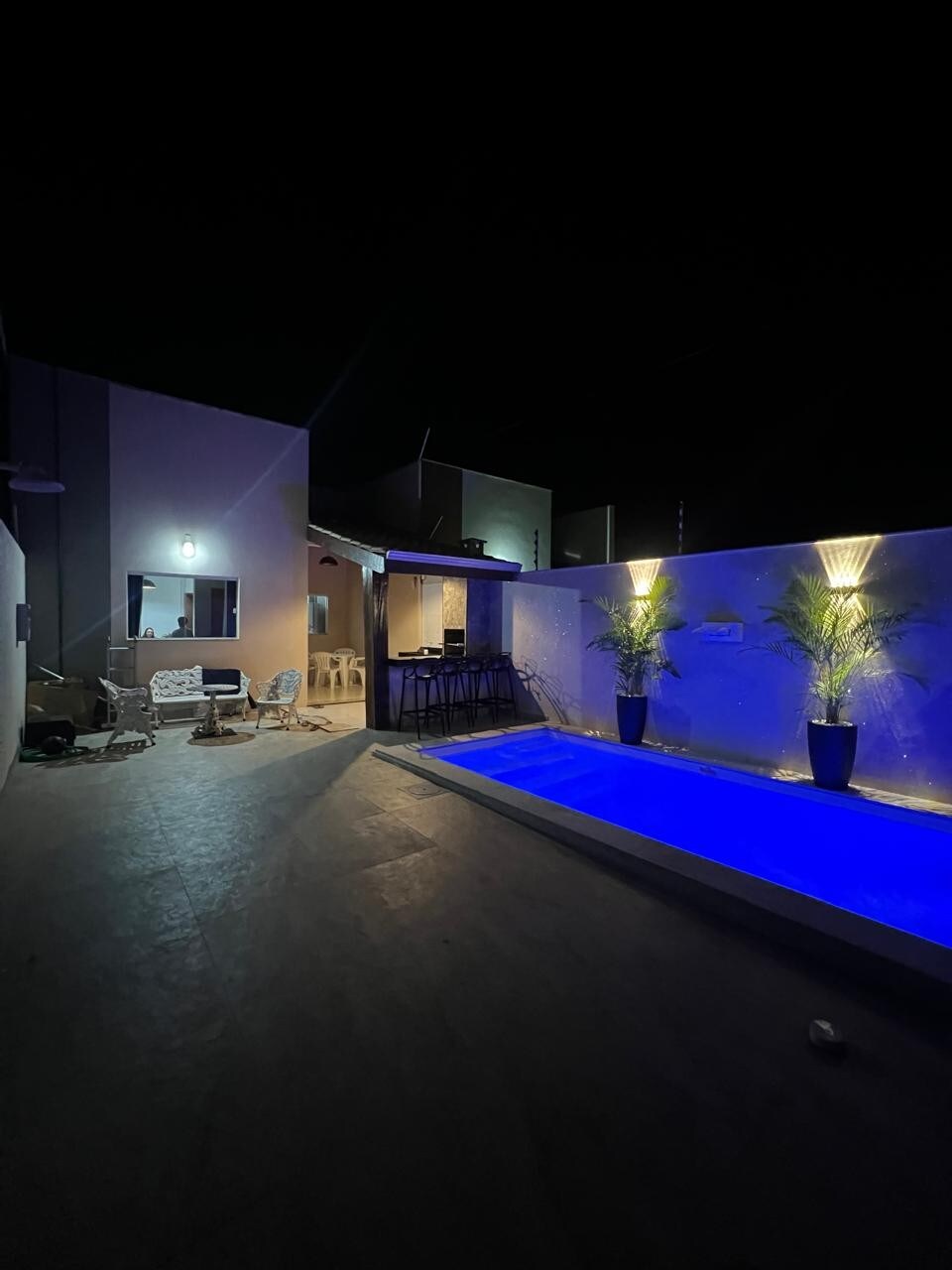 An outdoor area is illuminated by soft lighting at night, featuring a tranquil pool with blue water. Comfortable seating arrangements are positioned nearby, alongside tall plants in pots that add a touch of greenery.