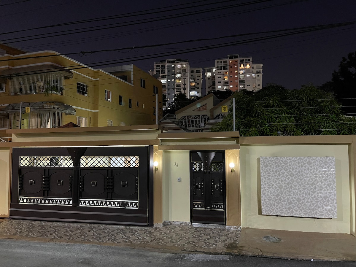 The exterior of a luxurious house is illuminated at night, showcasing a contemporary facade with decorative elements. A large double-gate entrance is visible, flanked by a solid door adorned with intricate designs. The urban skyline can be seen in the background, adding context to the setting.