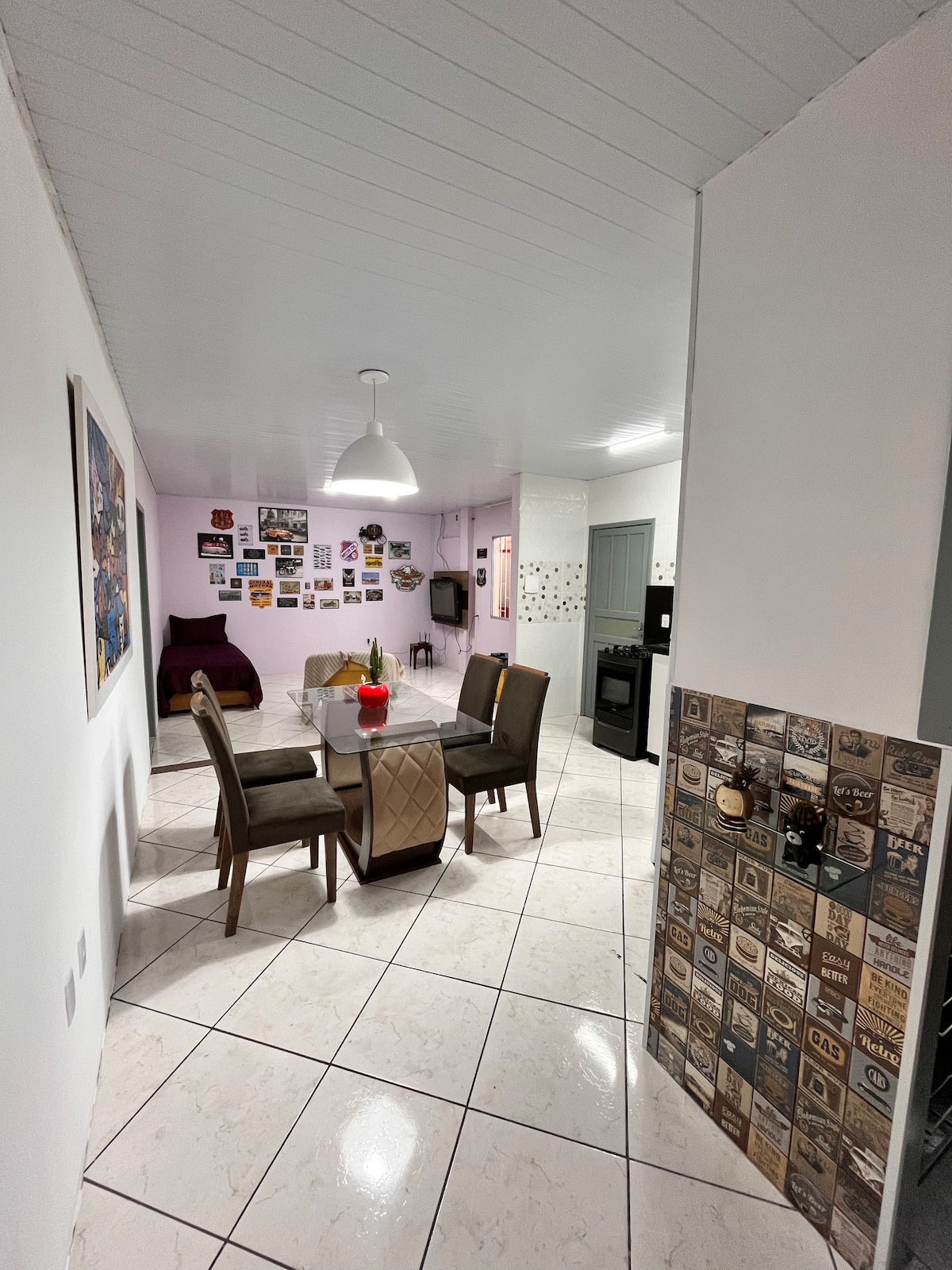 A bright and airy dining area features a modern glass table surrounded by four chairs. Neutral-toned walls are complemented by a variety of framed photographs. A cozy seating area with a purple chair is visible in the background, with a sleek, contemporary pendant light overhead.