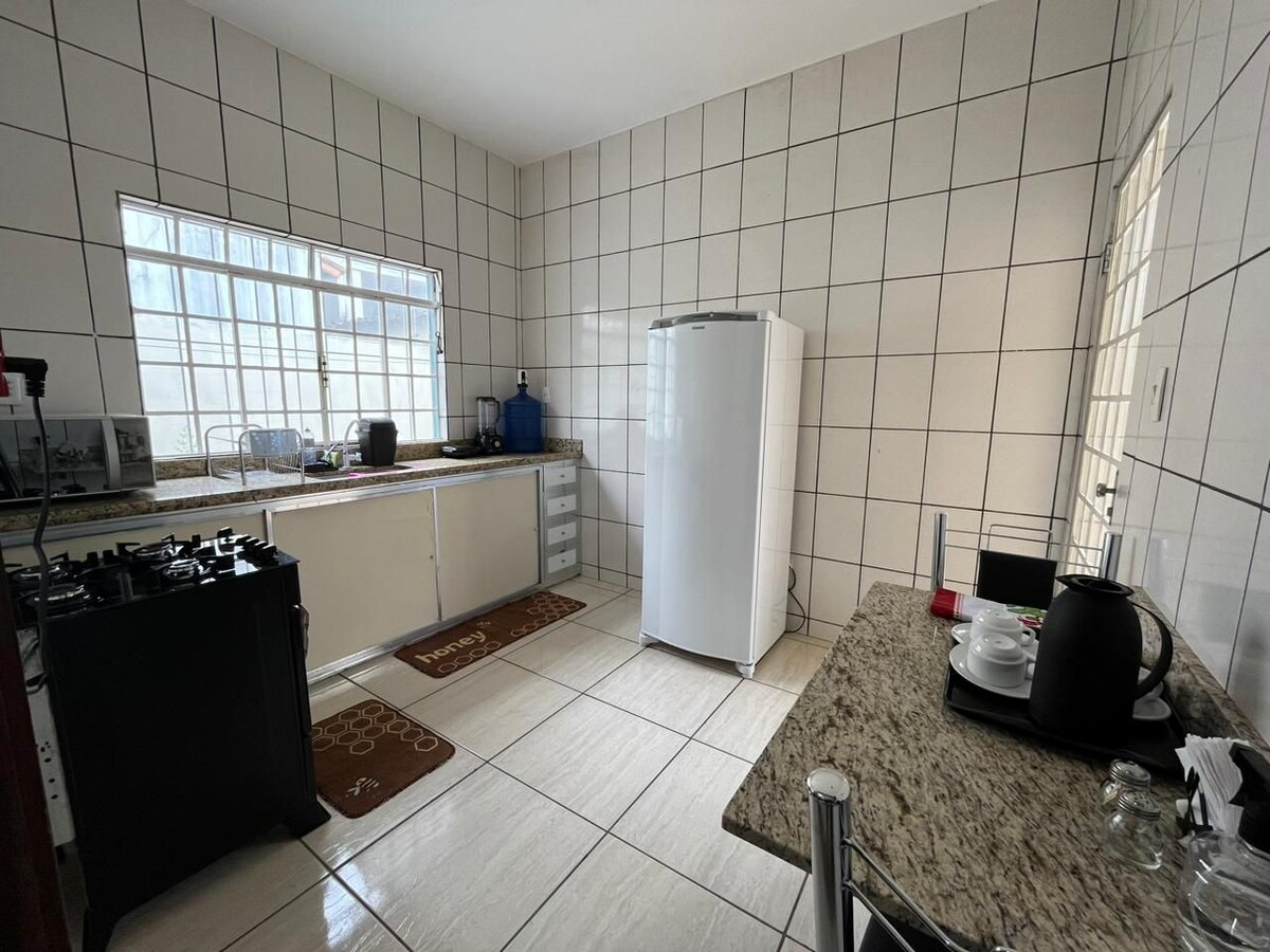 A spacious kitchen features tiled walls and a large window allowing natural light. A white refrigerator and a gas stove are present, along with a granite countertop. A small dining table is set with a kettle and glassware, enhancing the functional, welcoming environment.