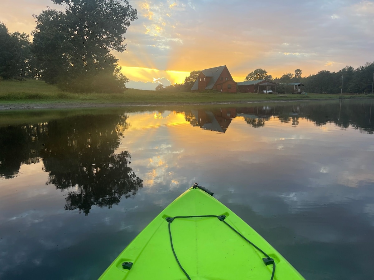 A vibrant green kayak is positioned in calm water, reflecting a stunning sunset with hues of orange and yellow. The background features a rustic cabin, subtly integrated into the natural landscape, surrounded by trees and open fields.