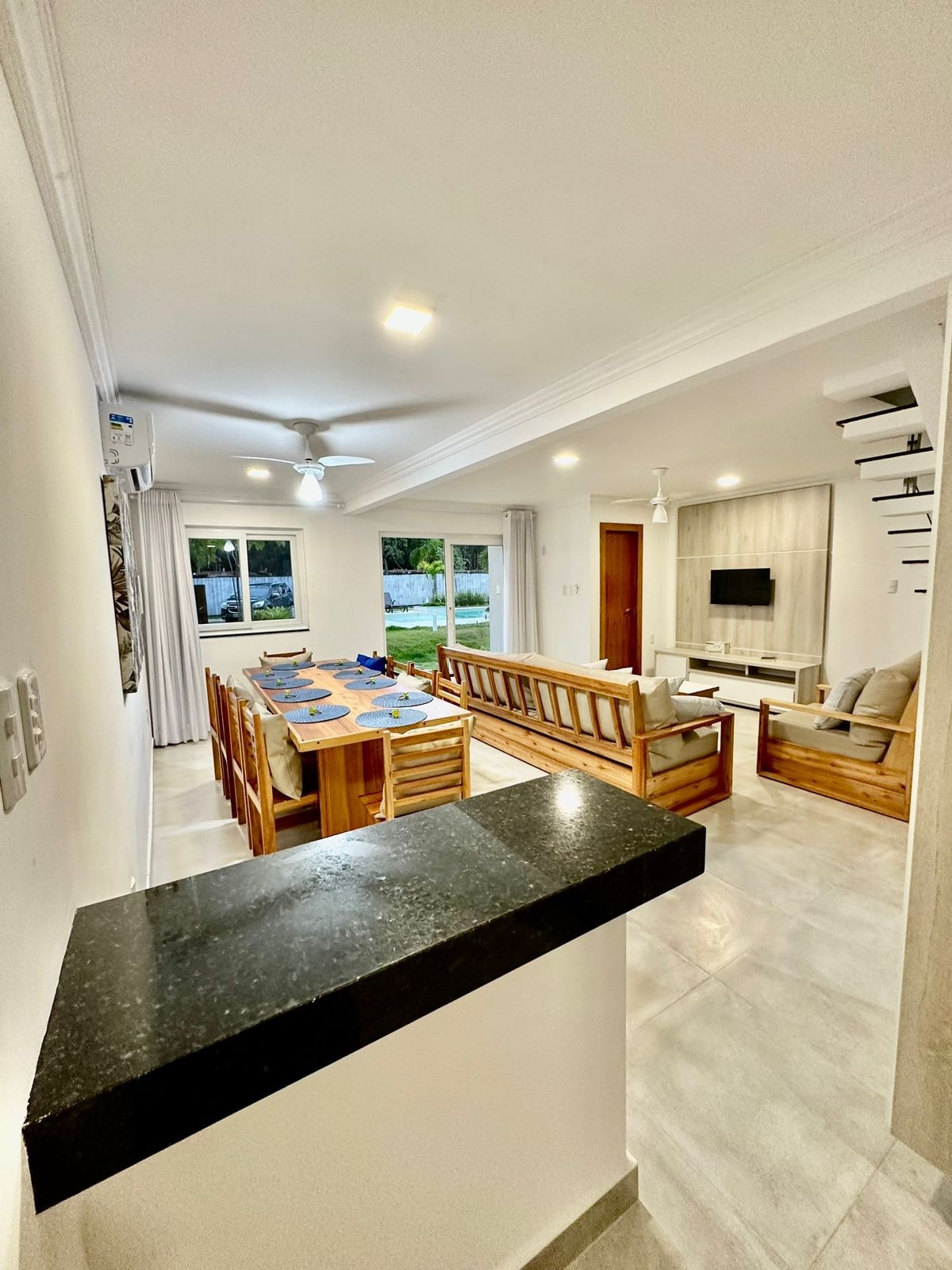 A bright living and dining area is shown, featuring a wooden dining table set for multiple guests. Comfortable seating is arranged in the living room, with light-colored furniture and large windows allowing natural light. A ceiling fan is visible above, enhancing the open feel.