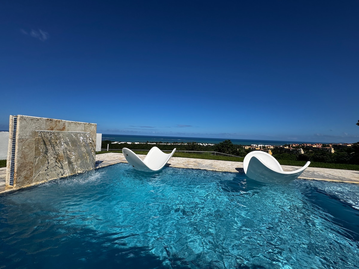 The private pool features two elegant loungers positioned on the water's edge. A gentle waterfall cascades from a stone wall, creating a serene atmosphere. The expansive view of the Atlantic Ocean is visible under a clear blue sky.