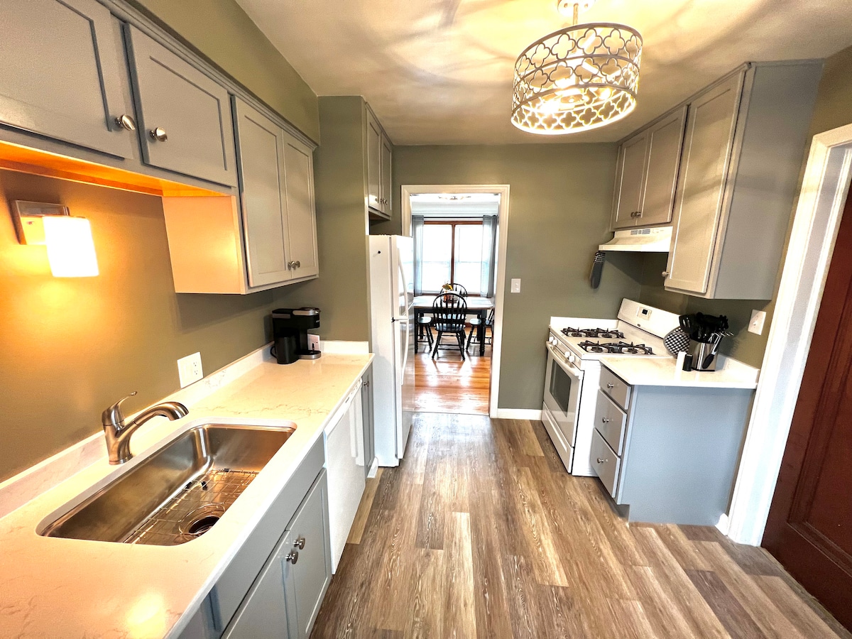 A newly renovated kitchen is presented with modern grey cabinetry and light countertops. Appliances include a refrigerator, stove, and coffee maker. Natural light filters through a nearby window, highlighting the open layout towards a dining area in the background.