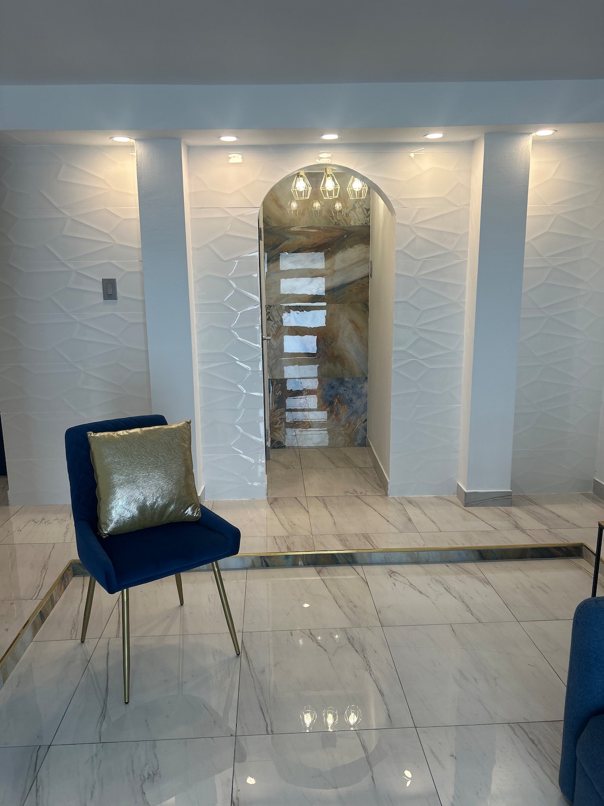 An elegant entryway leads into the apartment, featuring textured white walls and a marble-like floor. Soft lighting highlights the archway above, while a blue chair with a decorative gold pillow is positioned in the foreground, creating a welcoming atmosphere.