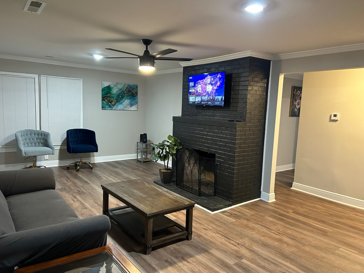 A spacious living room is highlighted by a dark brick fireplace, with a television mounted above it. Comfortable seating includes a gray sofa and two accent chairs, while a coffee table occupies the center space. Natural light enters through white-windowed blinds.