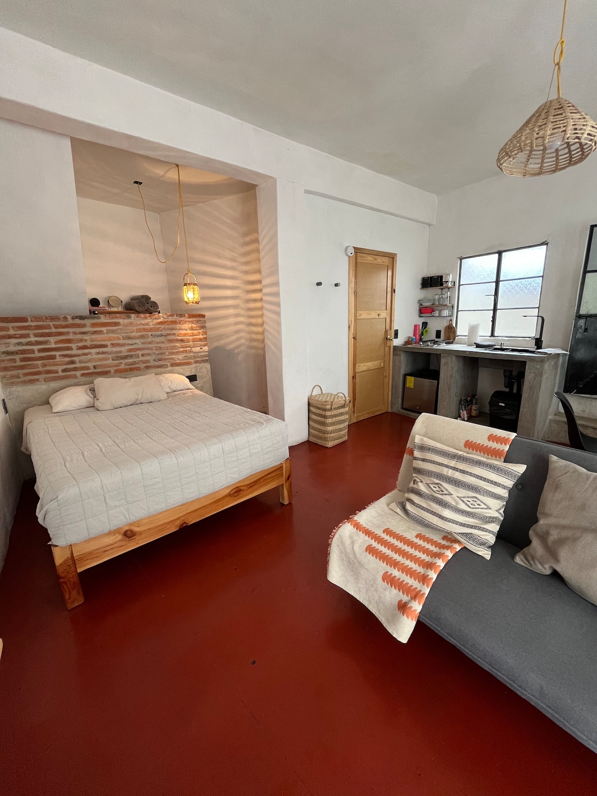 A cozy room features a spacious bed with a light-colored blanket, complemented by a decorative accent wall of exposed bricks. A small seating area is present, with a grey sofa adorned with a patterned throw. Natural light enters through a window, illuminating the interior.