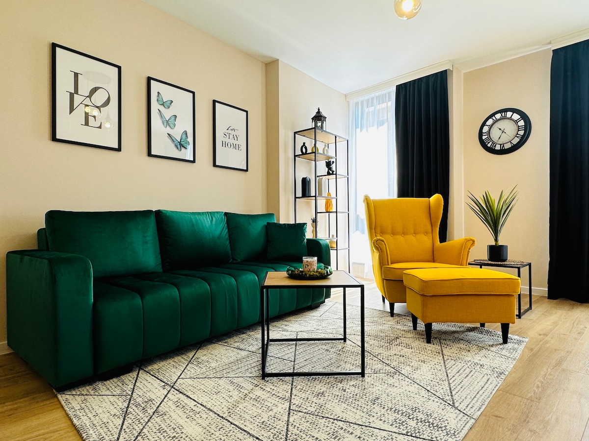 A modern living space features a green sofa and a yellow armchair, complemented by a coffee table. Artwork with butterflies and motivational phrases adorns the wall. A decorative shelf displays various items, while natural light streams in through a window, enhancing the cozy ambiance.