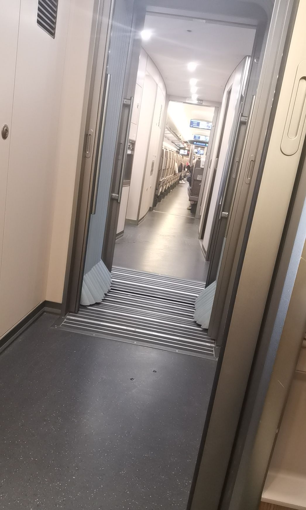 A narrow corridor is visible, featuring smooth flooring and segmented walls. Light fixtures illuminate the space, leading to a row of compartments on both sides. The layout suggests convenience for navigating between areas within the building.