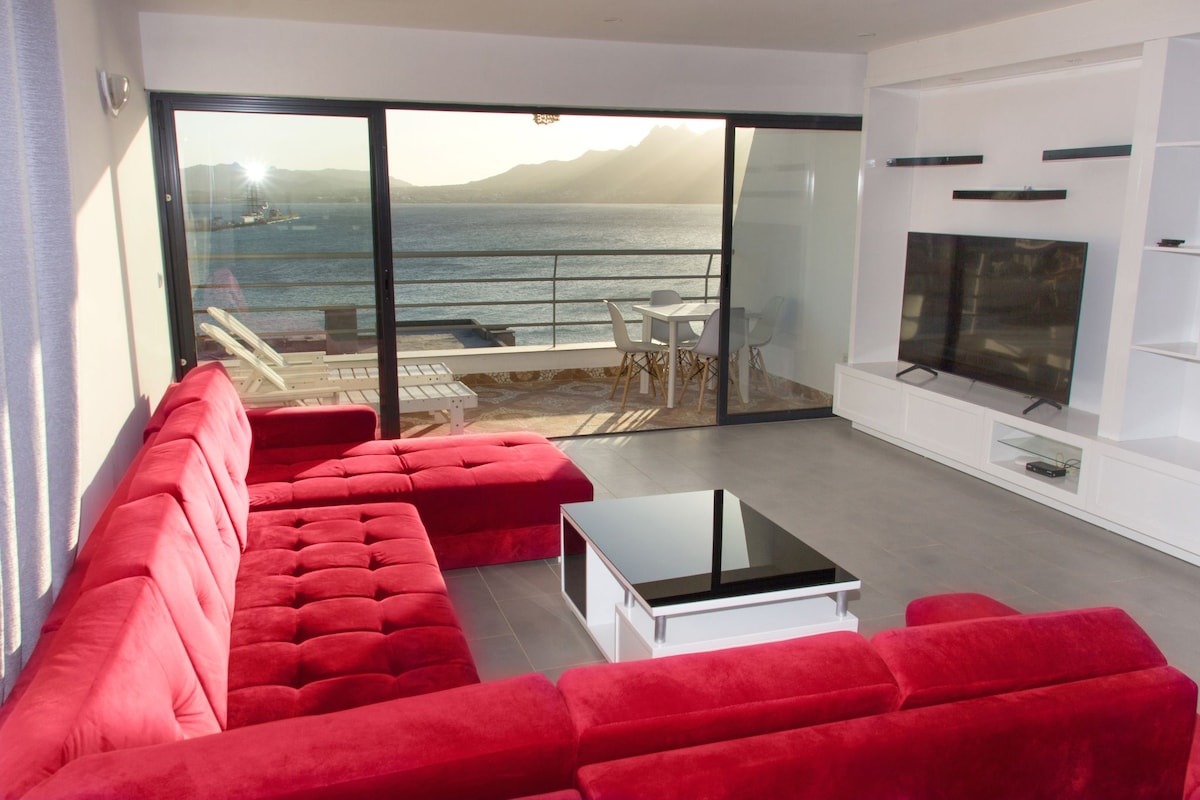 A spacious living area features a large red sectional sofa surrounding a coffee table. Floor-to-ceiling glass doors offer a view of the ocean and terrace. A television is mounted on the wall, and a dining area with a table and chairs is visible in the background.