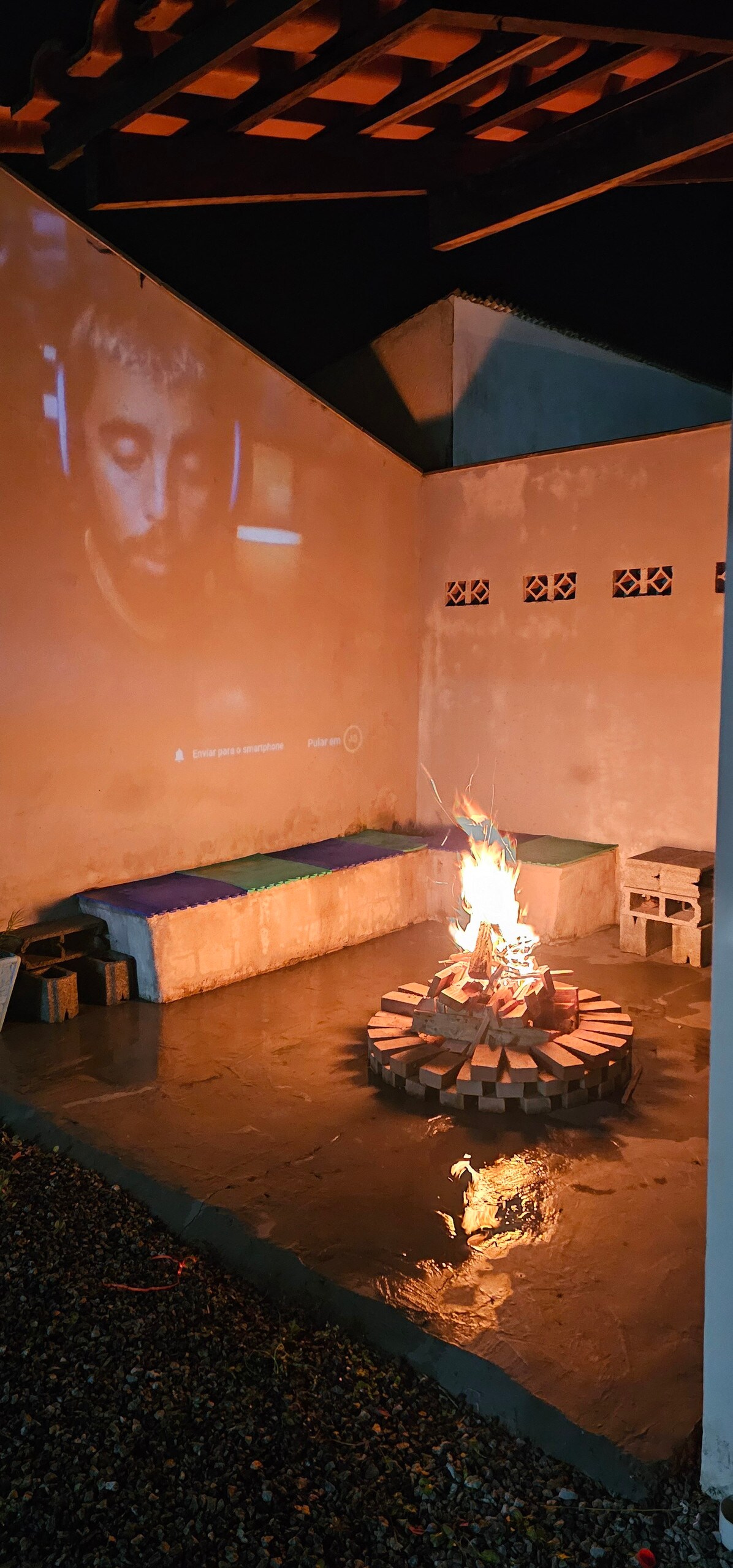 An outdoor seating area is featured, highlighted by a circular fire pit surrounded by logs. A projection screen is visible on one wall. Blankets are arranged on the seating, which offers a cozy spot to gather. The surface is damp, indicating recent weather.