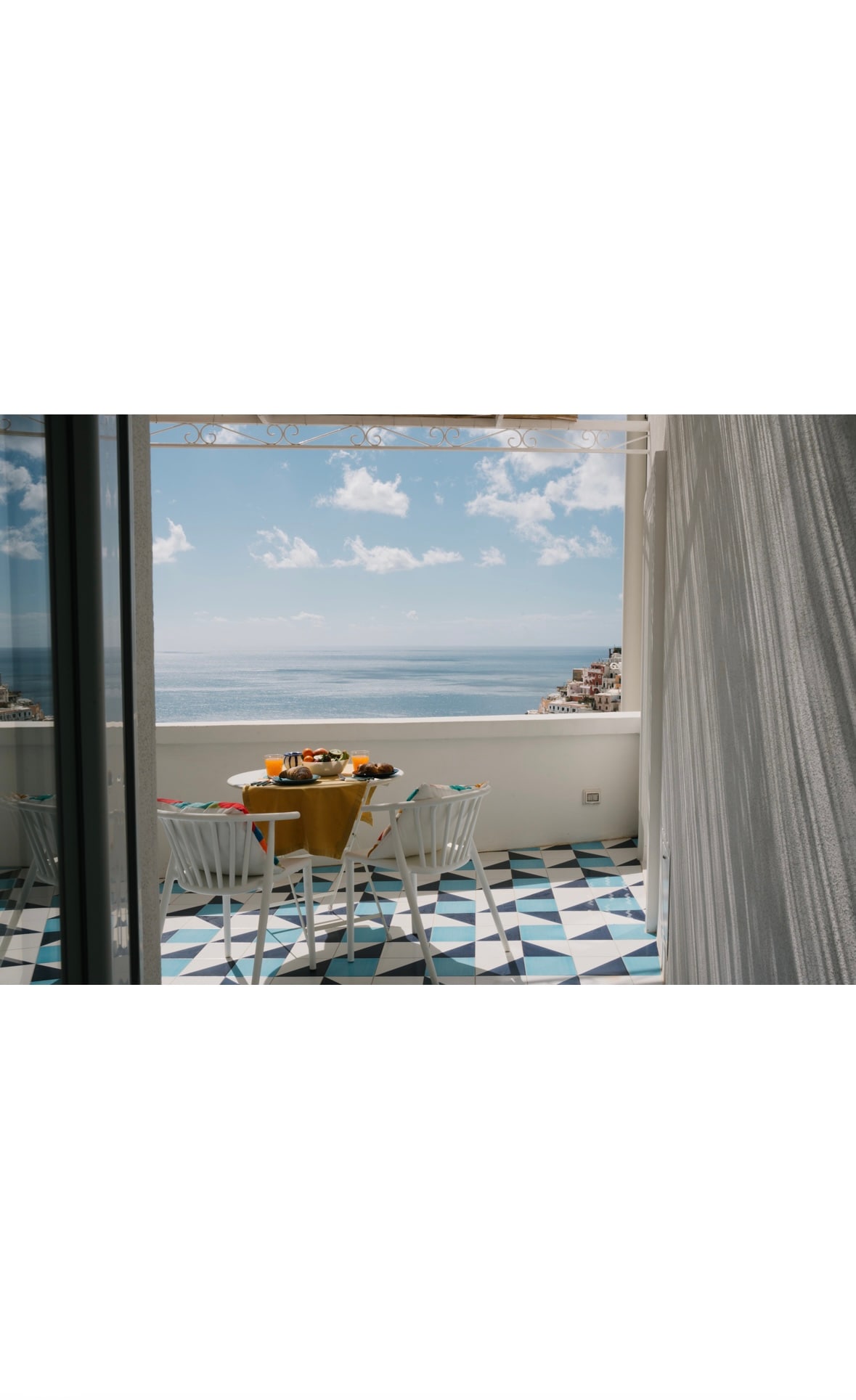 A serene terrace features a round table set for breakfast, accompanied by four white chairs. The stunning view of the sea is framed by the bright blue sky adorned with fluffy clouds. The patterned floor tiles add a vibrant touch to the outdoor space.
