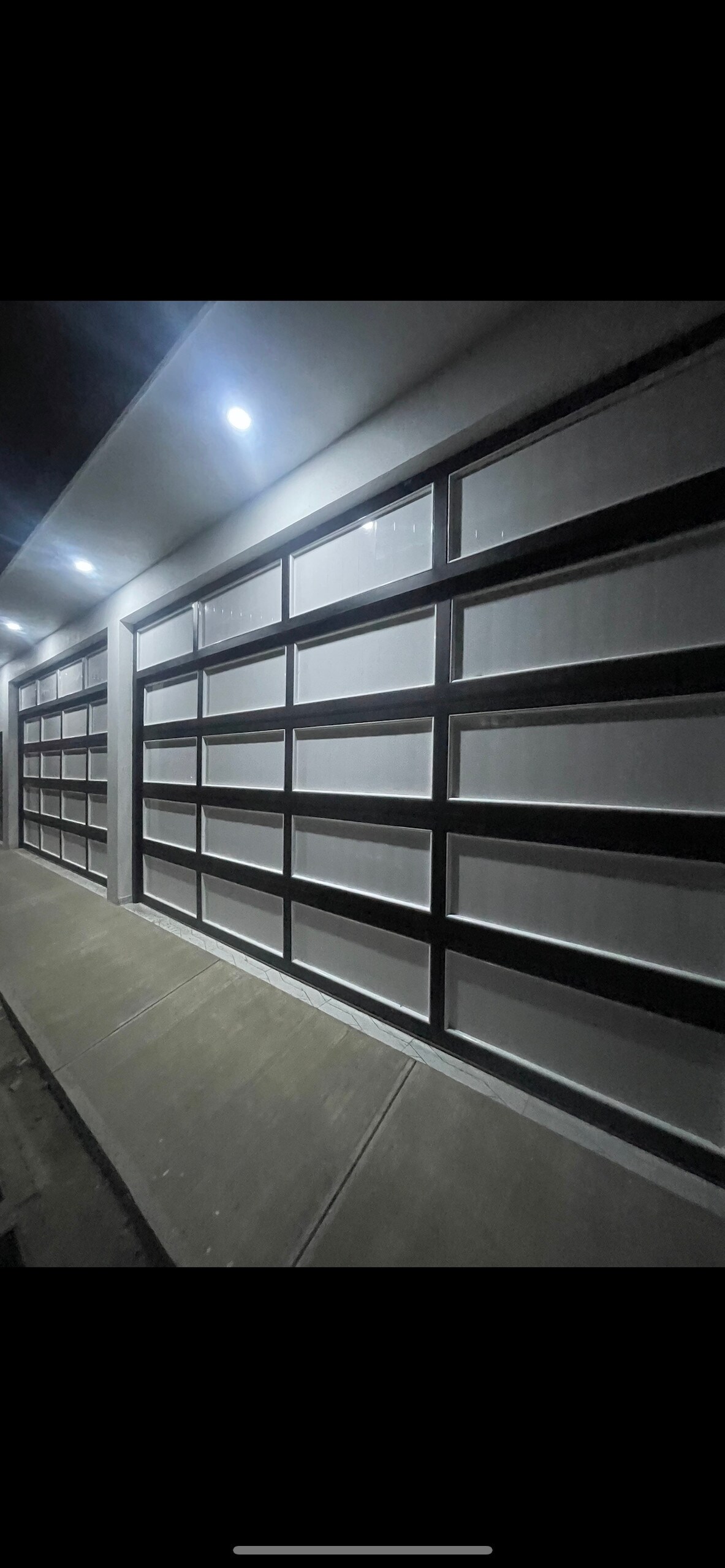 A modern garage facade is shown, featuring multiple large glass panels framed with black metal. The sleek design is illuminated by overhead lights, enhancing the contemporary aesthetic. Clean lines and a minimalist approach contribute to the overall appeal of the space.