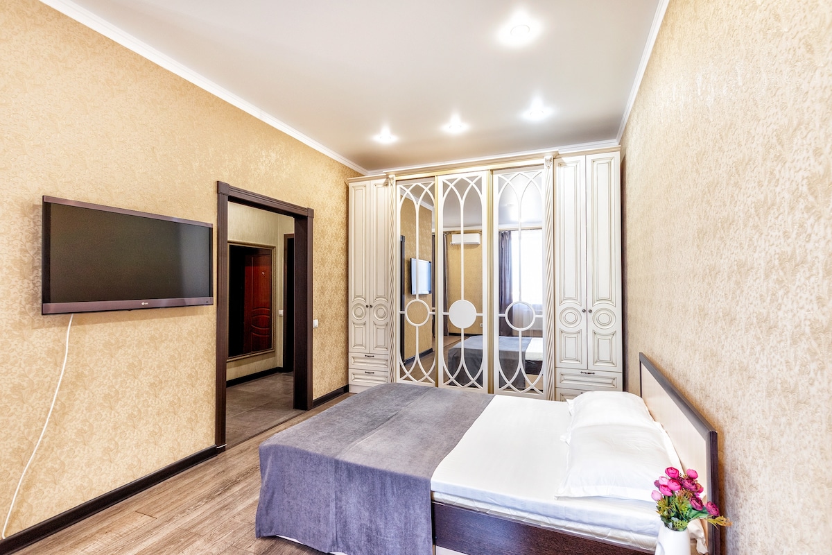 A soothing bedroom features a spacious bed covered with a soft blanket. A wall-mounted television is displayed opposite the bed. Mirrored wardrobes with decorative detailing line one wall, enhancing the room's elegance. Soft lighting from recessed fixtures provides a warm ambiance.