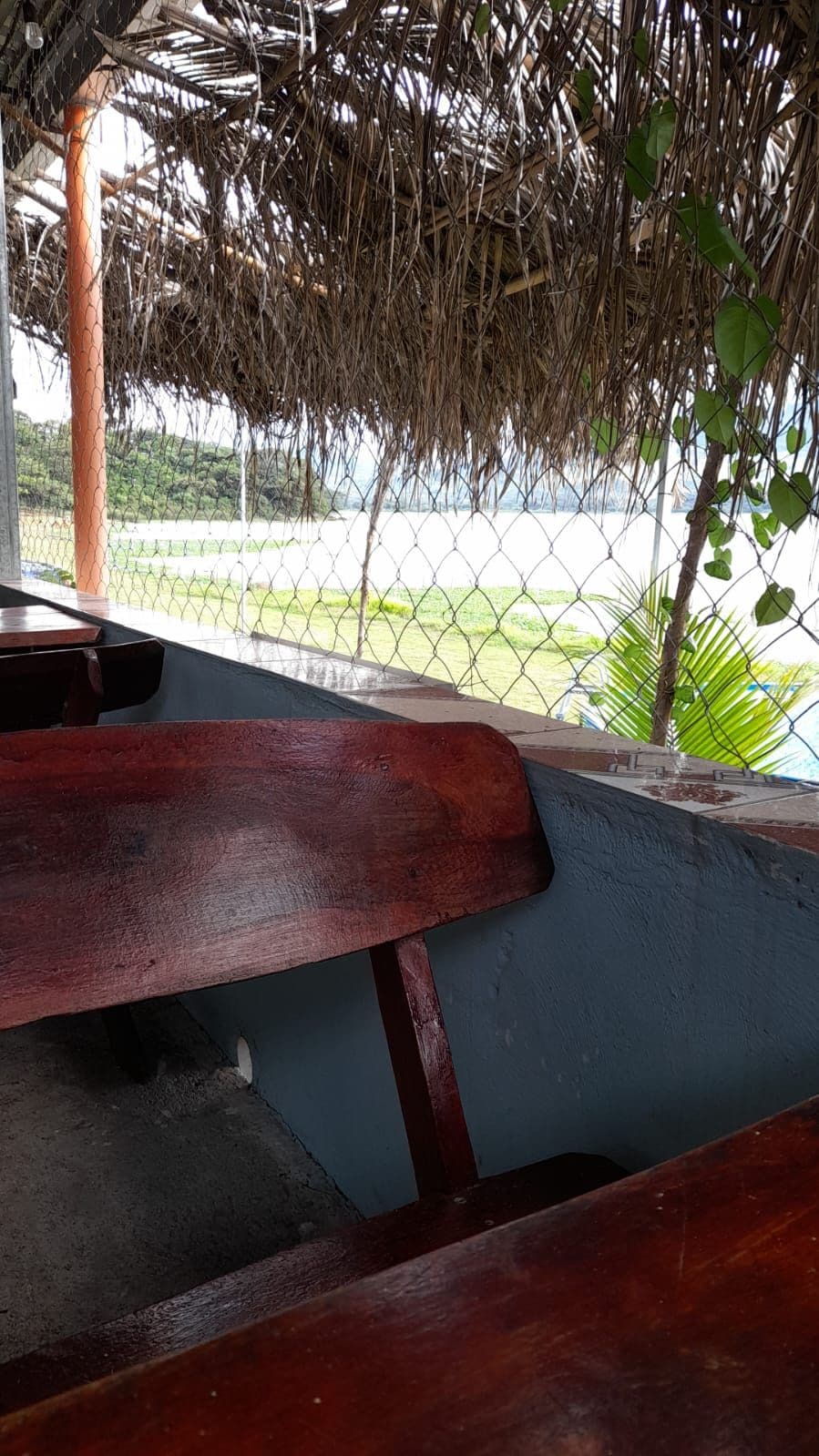 Wooden benches are positioned against a stone wall, offering a relaxed seating area. A thatched roof provides shade, while a view of open green space and water is visible in the distance, framed by a fence and palm leaves.