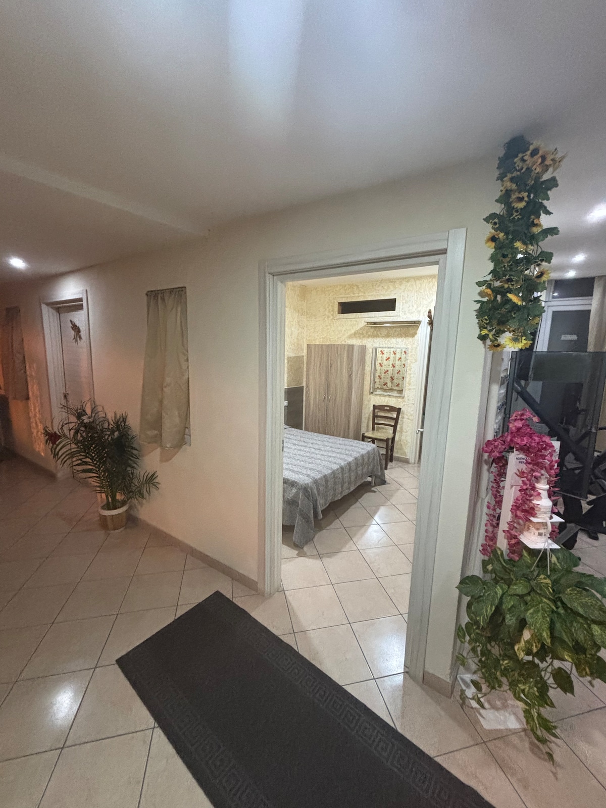 A hallway leads to a cozy bedroom, featuring a neatly made bed with a gray patterned coverlet. Decorative plants and floral arrangements adorn the area, while soft lighting creates a welcoming ambiance. Walls are accented with light tones, enhancing the overall space.