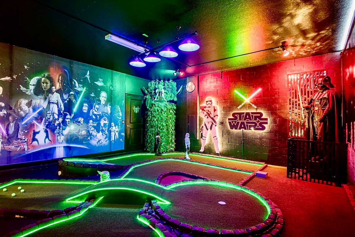 A themed indoor mini-golf area features vibrant Star Wars artwork on walls illuminated by colorful lights. Green neon outlines define the putting course, while decorative elements resemble plants and characters, creating an engaging atmosphere for families.