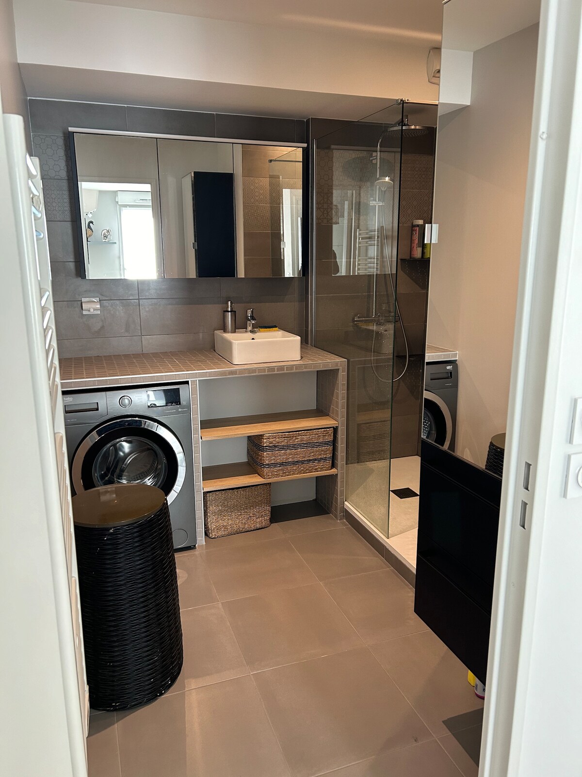 A modern bathroom features a sleek shower enclosure with a clear glass door. A wash basin is positioned on a countertop, alongside a stacked washer and dryer. Shelving units provide additional storage, and a black wicker basket adds a textured element to the space.