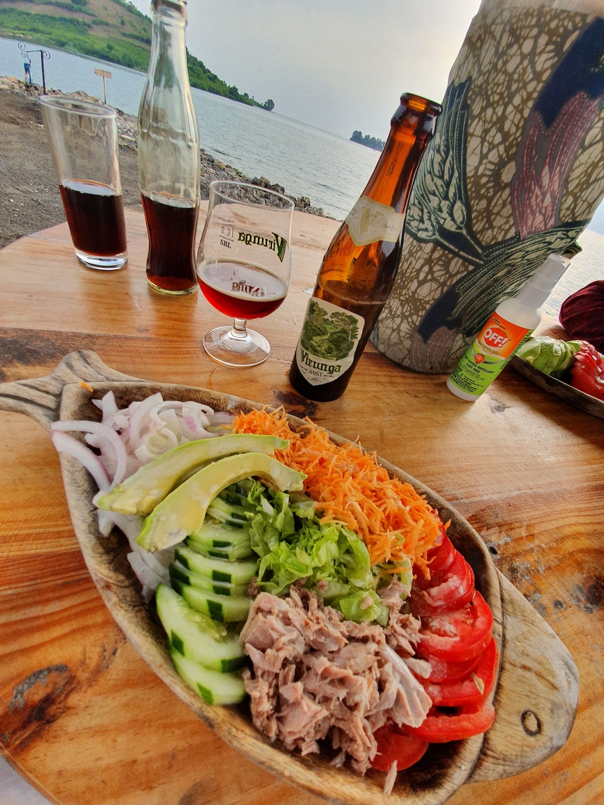 A wooden table holds a vibrant platter of fresh vegetables and tuna, arranged with slices of cucumber, tomato, and avocado. Nearby, two glasses of local beverages are present, along with a beer bottle and a bottle of insect repellent, set against a backdrop of water and greenery.