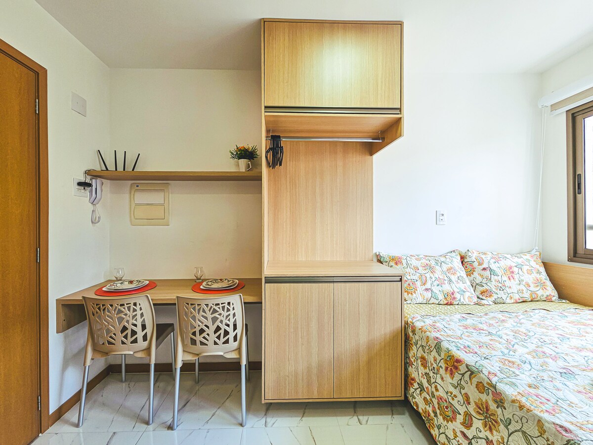 A compact studio living space is highlighted by a comfortable bed adorned with a floral-patterned bedspread. A dining area features two chairs and a table, with a wall-mounted shelf above. Warm wood cabinetry complements the light-colored walls and window, providing natural light.