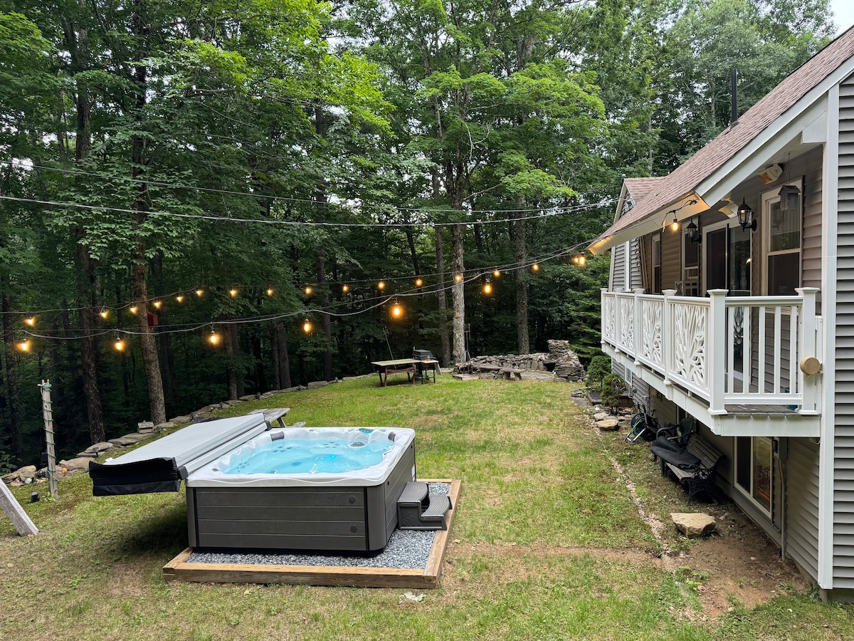 A hot tub is positioned on a wooden deck in the backyard, surrounded by trees and soft lighting from overhead string lights. A grassy area is present nearby, along with a picnic table and stone features, providing a serene outdoor space.