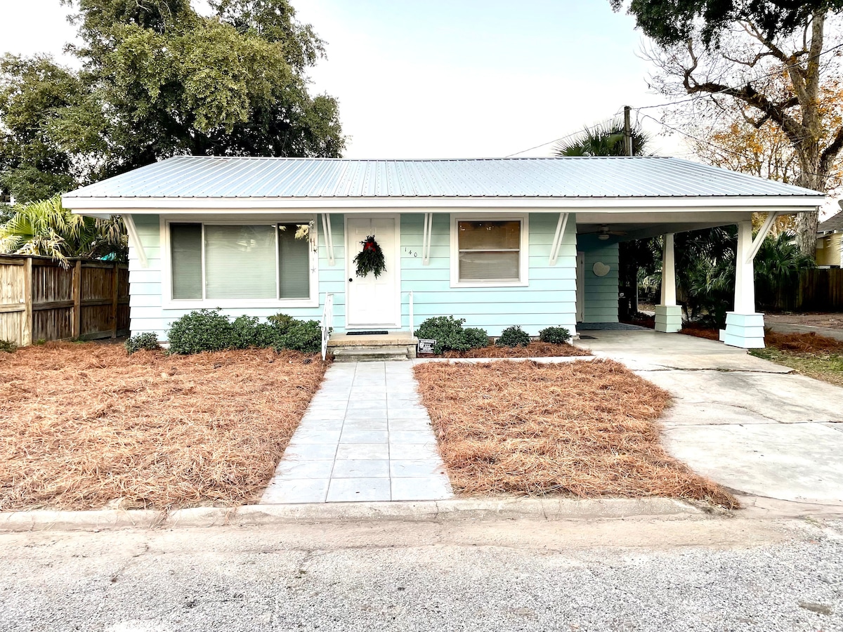 A charming blue house with a metal roof is situated on a quiet street. A walkway leads to the front door, bordered by neat landscaping and shrubs. The yard is covered with freshly raked pine straw, creating an inviting entrance.