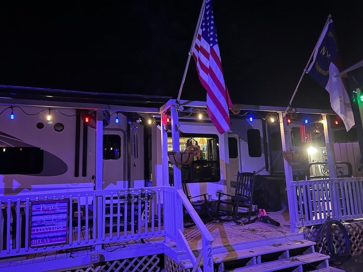 A charming exterior view of a trailer at night, adorned with colorful lights. An American flag is displayed prominently alongside another flag. Rocking chairs provide seating on the front porch, enhancing the welcoming atmosphere.