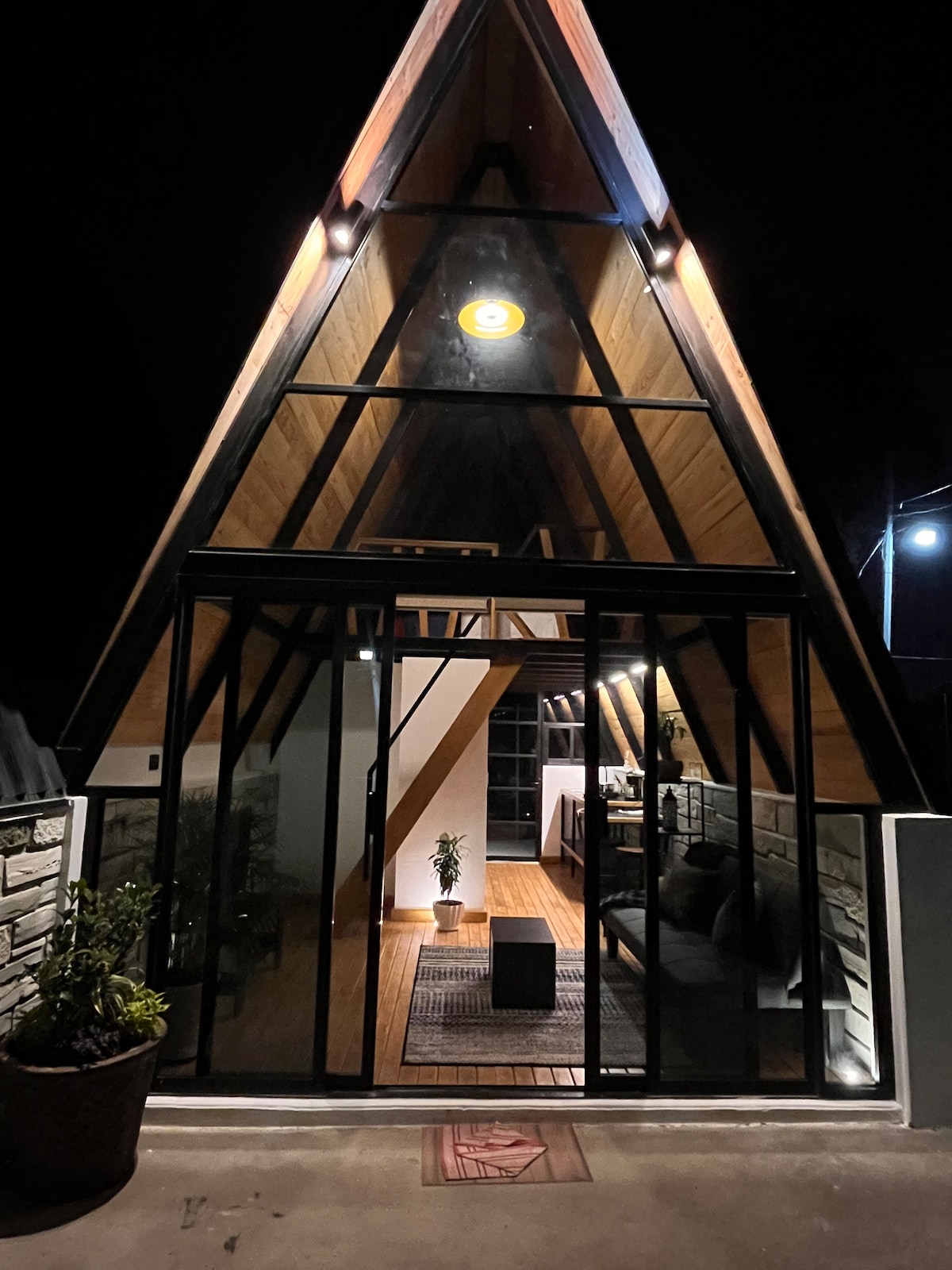 The A-frame cabin is showcased in an evening setting, with large glass doors providing a view of the interior. Warm lighting illuminates wooden accents and a spacious layout, enhancing the cozy atmosphere. A small potted plant is visible near the entrance.