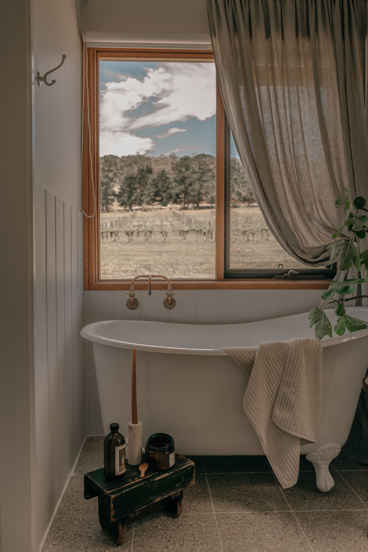 A serene bathroom features a freestanding tub beside a large window offering a view of the vineyards. Natural light filters through sheer curtains, enhancing the calming atmosphere. A small stool holds artisan toiletries, while a soft blanket drapes casually over the tub's edge.