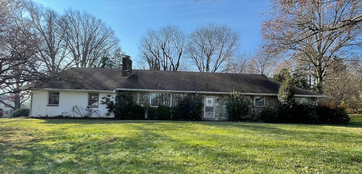 A single-story ranch house is set against a clear sky, surrounded by a spacious green lawn and mature trees. Large windows enhance the bright exterior, while a central entrance provides access to the home. Lush landscaping completes the inviting front yard.