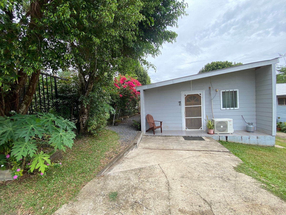 A welcoming exterior of a bungalow is framed by vibrant greenery, including tropical plants and a flowering bush. A pathway leads to the entrance, which features a wooden chair on the patio beside the door. The facade displays a window and an air conditioning unit.