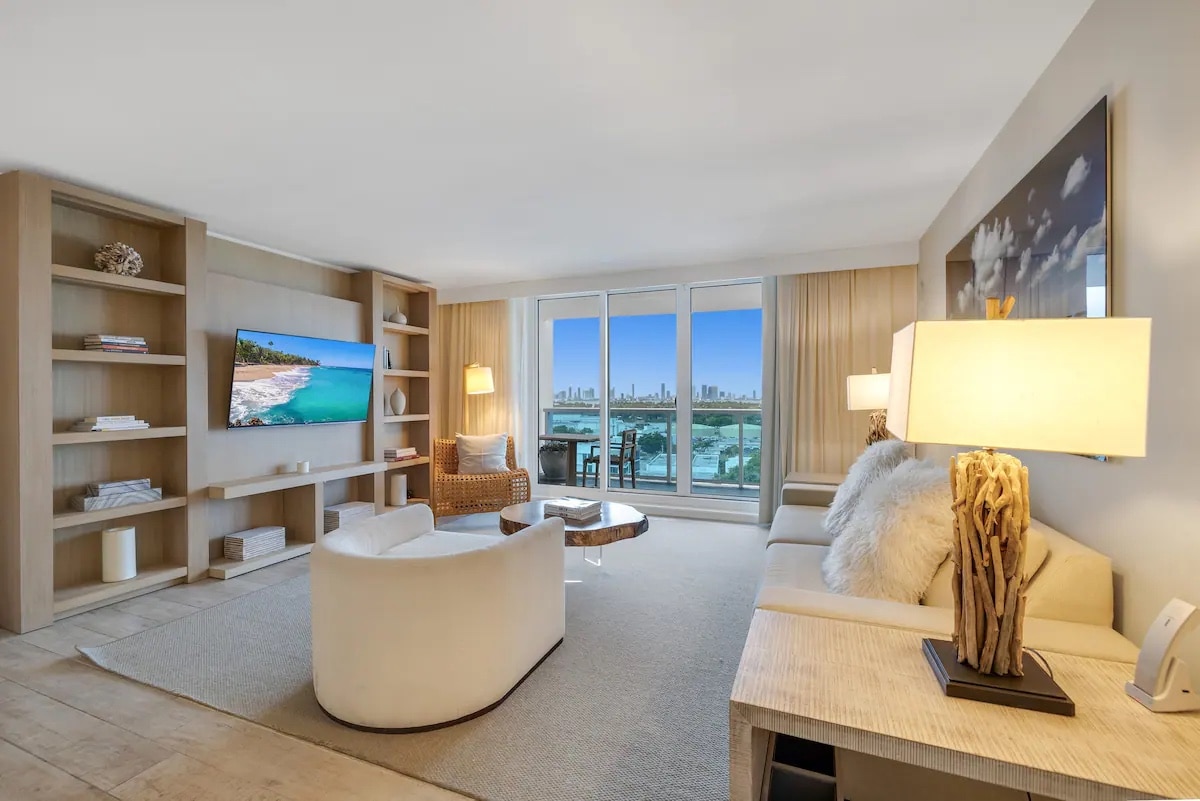 A stylish living area showcases a spacious seating arrangement, including a curved chair and a light-colored sofa. A flat-screen television is mounted on the wall, with a wooden shelf displaying decorative items. Large windows offer views of the outdoor scenery and natural light.