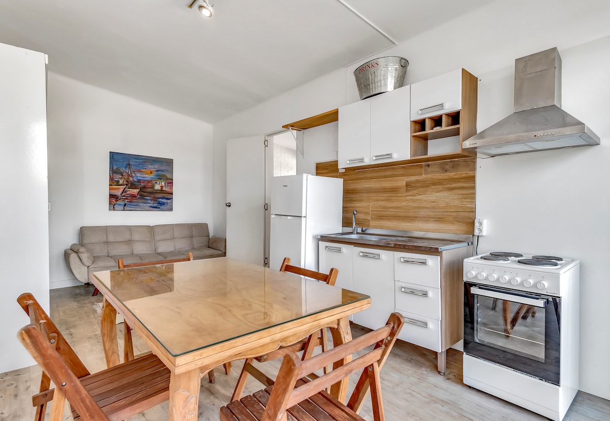 A functional kitchenette is visible with wooden cabinetry and stainless steel appliances. A dining table with wooden chairs is positioned centrally, and a cozy sofa sits against the wall. A colorful artwork adds a touch of character to the inviting space.