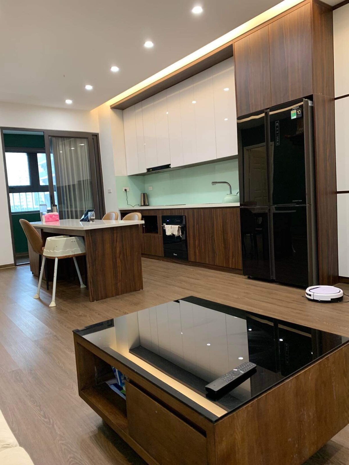 A modern kitchen features a combination of dark wood and glossy white cabinetry. Counter space is provided for meal preparation, alongside essential appliances including a refrigerator and dishwasher. A small dining area is located beside the counter, and a sleek coffee table is placed in the adjoining living space.