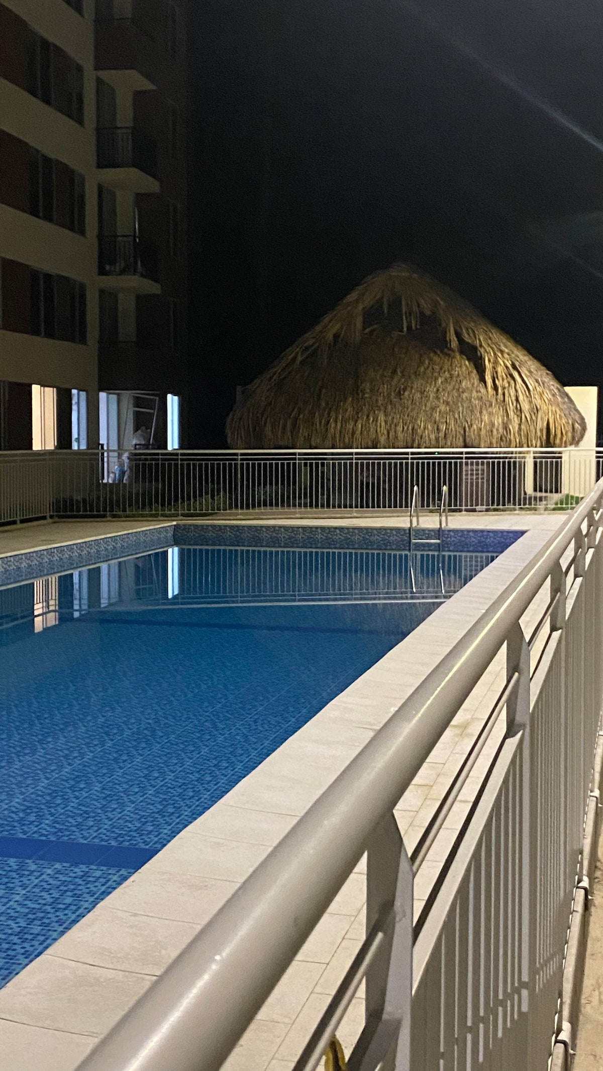 A serene pool area is illuminated at night, featuring a well-maintained swimming pool surrounded by a white railing. A thatched-roof structure is visible nearby, contributing to the relaxing ambiance of the outdoor space.