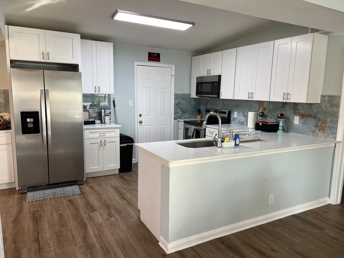 A modern kitchen is displayed featuring white cabinetry, stainless steel appliances, and a spacious island with a sink. Natural light illuminates the room, highlighting the light-colored counters and flooring. The kitchen is equipped with essential cooking items, ensuring convenience for meal preparation.