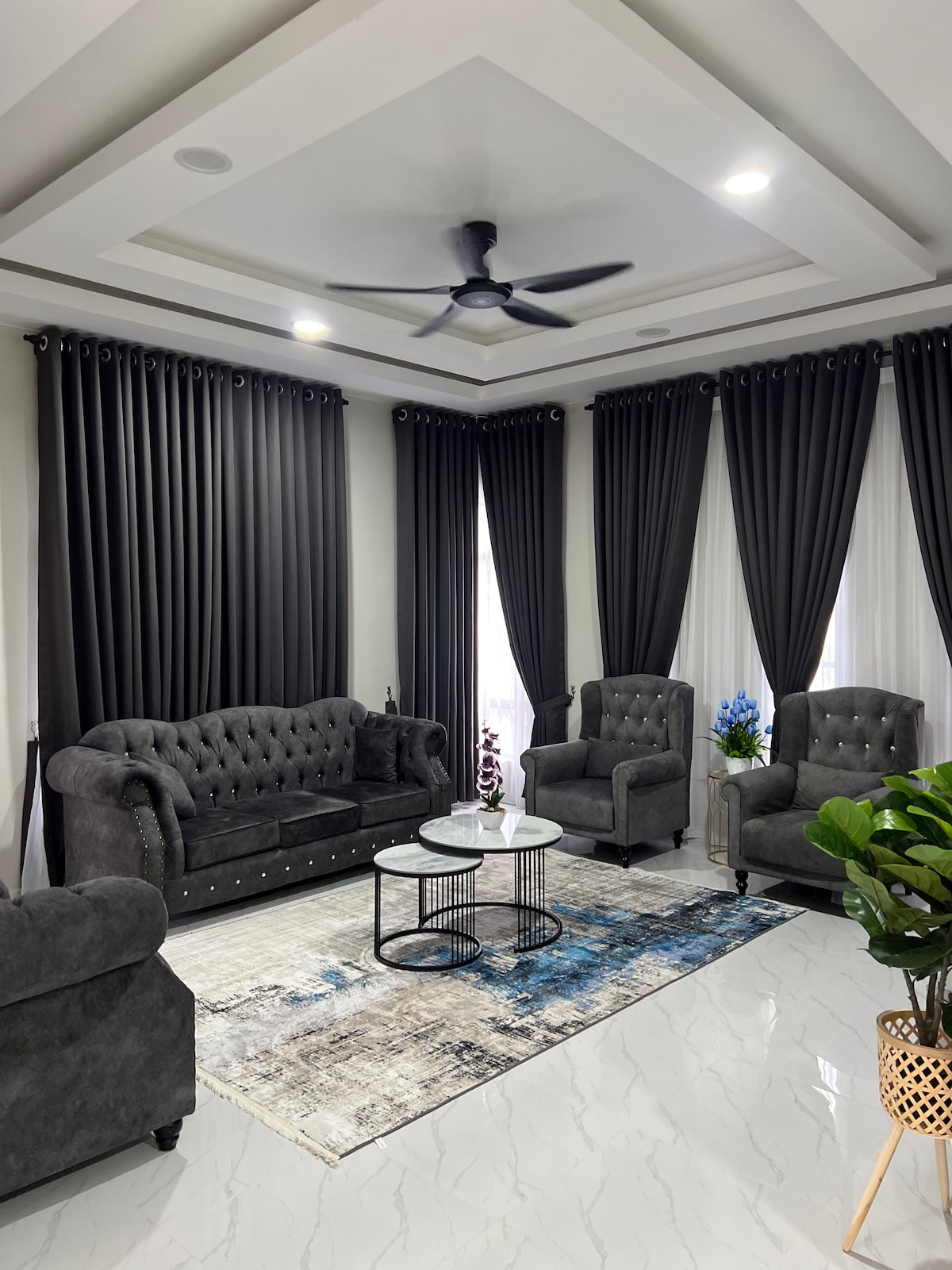 A living room is showcased with plush, dark grey sofas and armchairs arranged around a round coffee table. Elegant curtains frame the large windows, allowing soft light to illuminate the space. A decorative rug adds texture beneath the seating area, complemented by a vibrant plant in a corner.