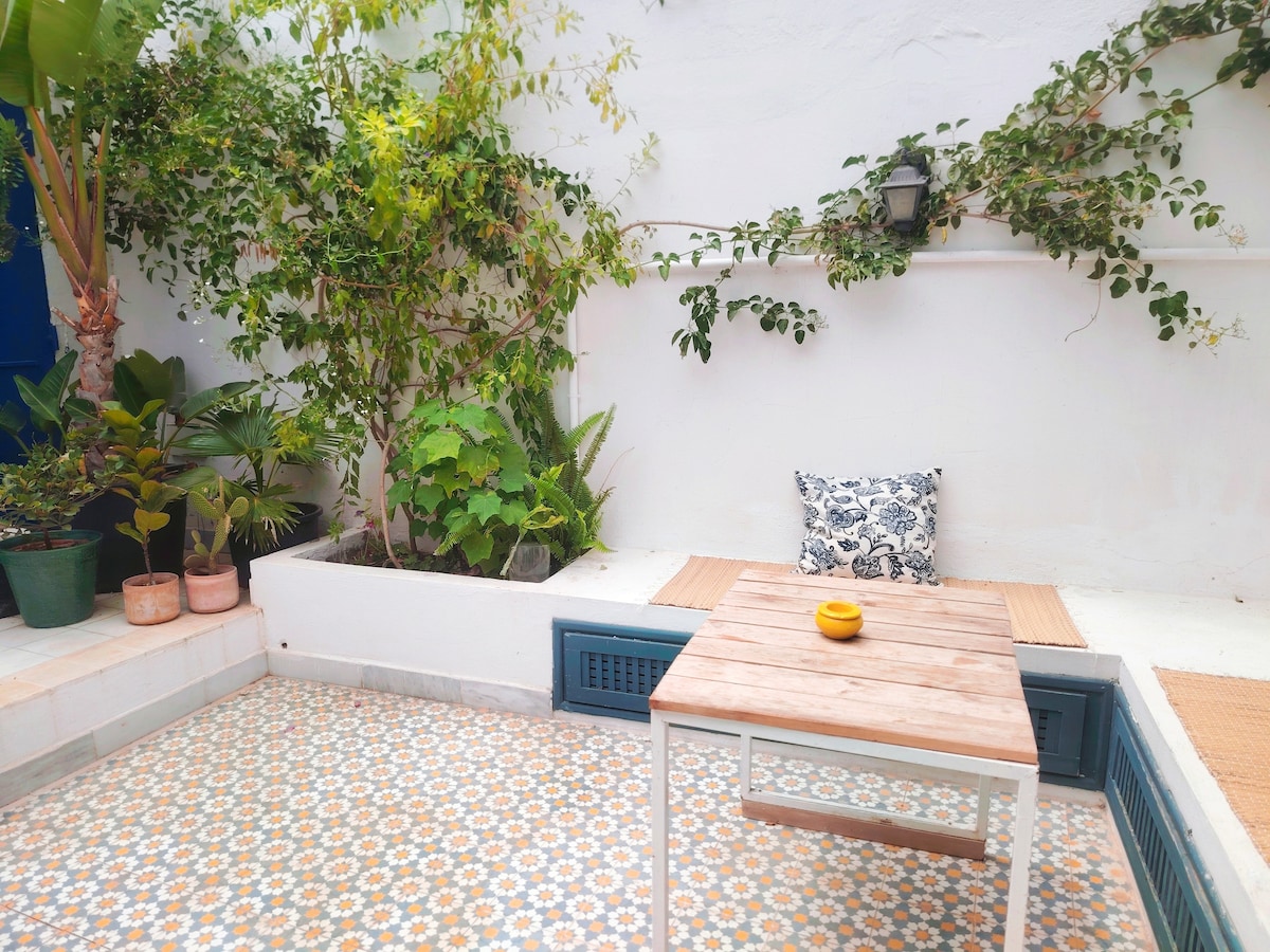 An outdoor terrace is shown, featuring a wooden table and a small yellow bowl. Surrounding plants provide greenery against the white wall, while patterned tiles add texture to the ground. A decorative cushion is visible on the seating area, enhancing the serene environment.