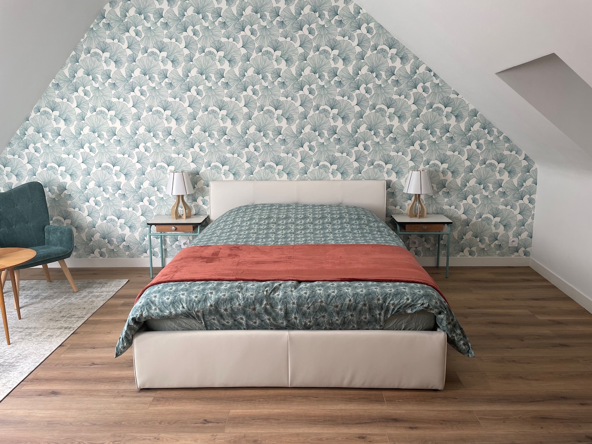 A spacious sleeping area features a large upholstered bed with a decorative coverlet. Two bedside lamps are positioned on teal tables. The wall is adorned with a floral wallpaper, complementing the light wooden flooring. A cozy seating area is visible to the side.