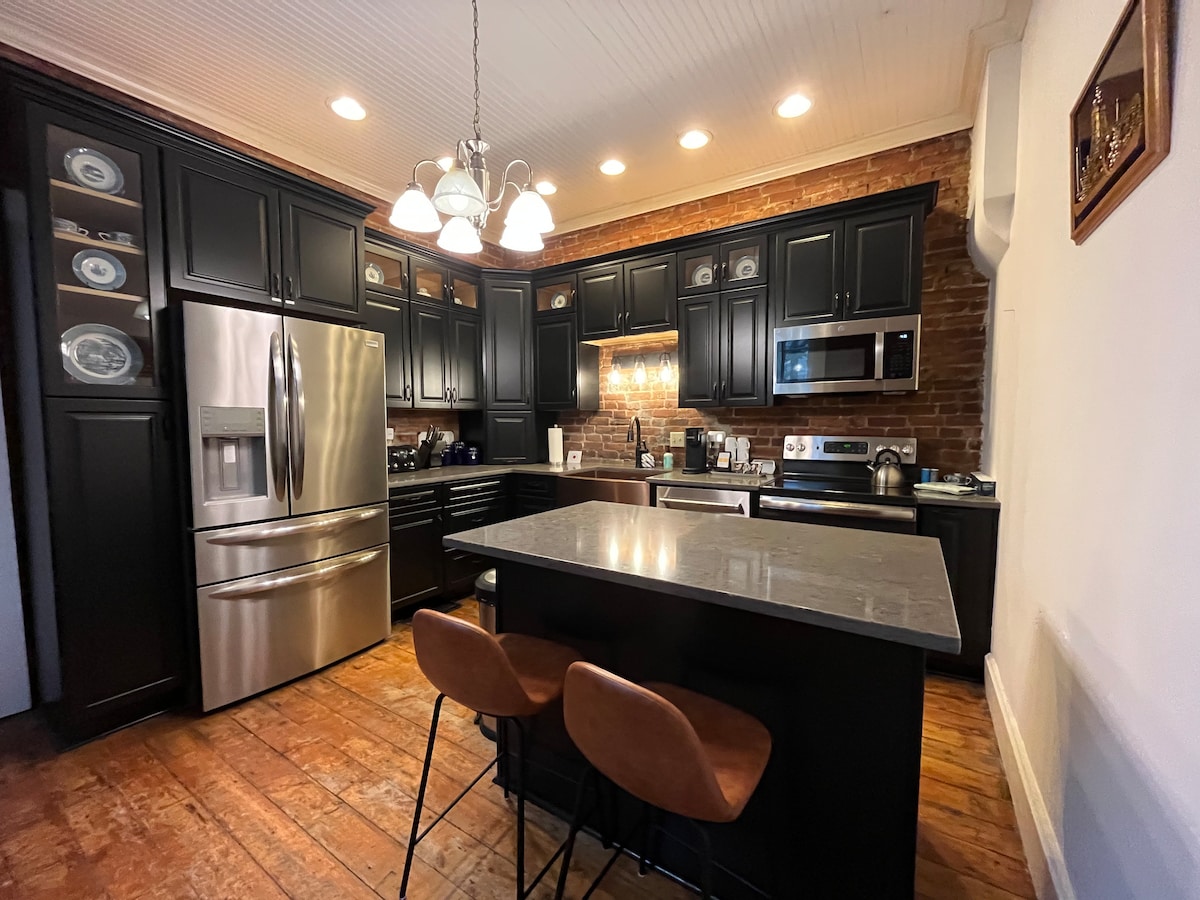 The kitchen features dark cabinetry paired with a light grey island countertop. Stainless steel appliances, including a refrigerator and microwave, are visible. A brick accent wall adds texture, and pendant lighting hangs above the island. Two bar stools provide seating.