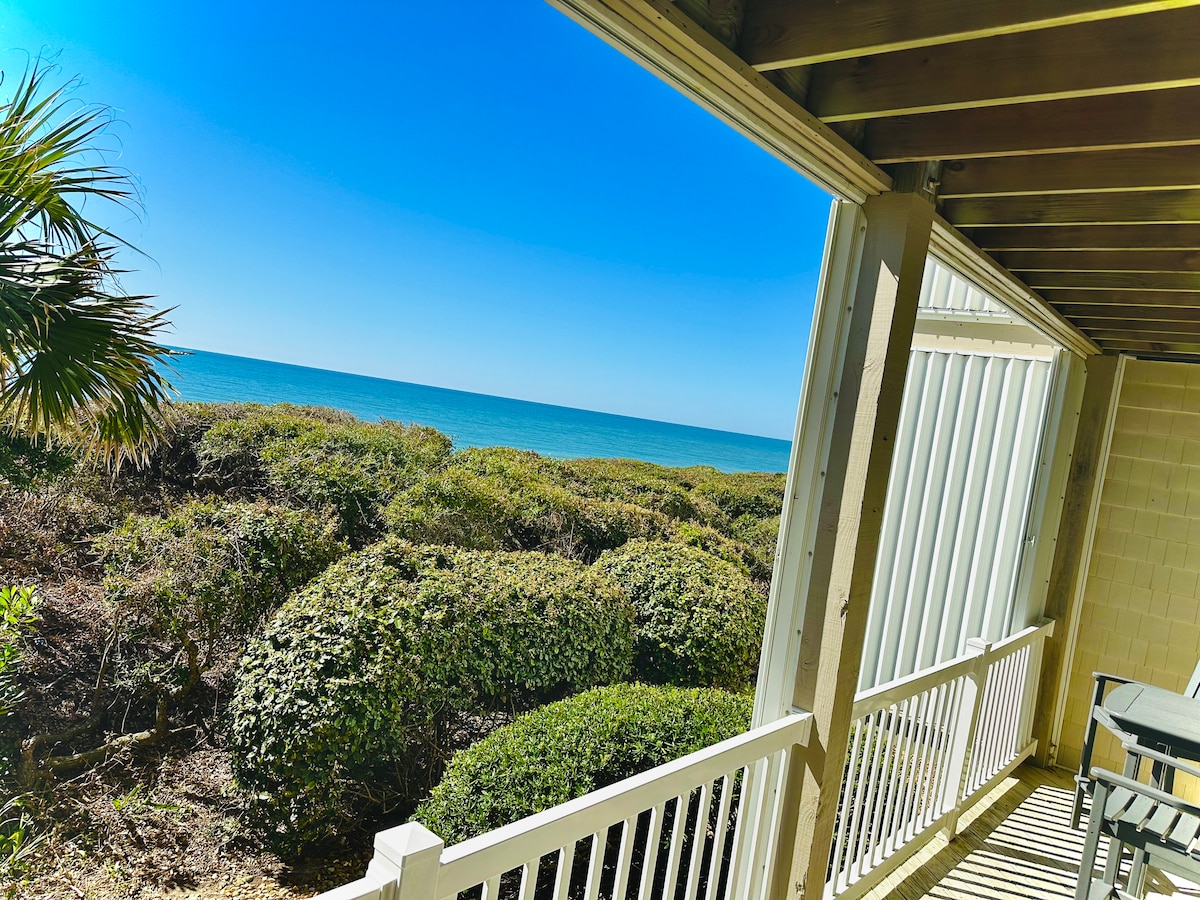 A spacious deck offers a clear view of the ocean, framed by lush green coastal vegetation. The sunlit space features slatted railings and provides an ideal spot to relax while enjoying the surrounding natural beauty.