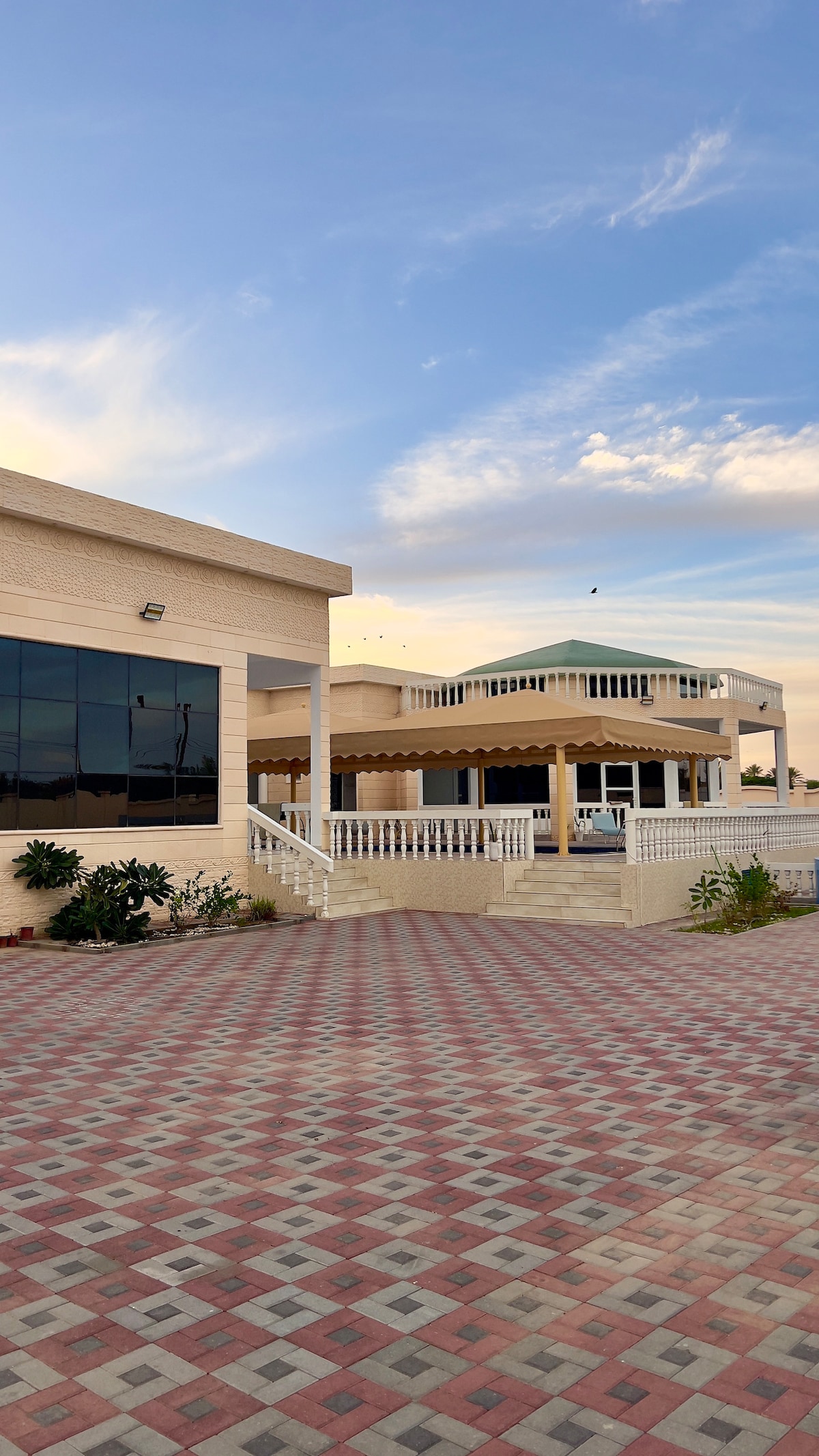 The exterior of the complex showcases a spacious layout with a patterned stone driveway. Two distinct architectural structures are visible, one featuring large windows and the other with a covered terrace, offering a welcoming entrance surrounded by greenery.