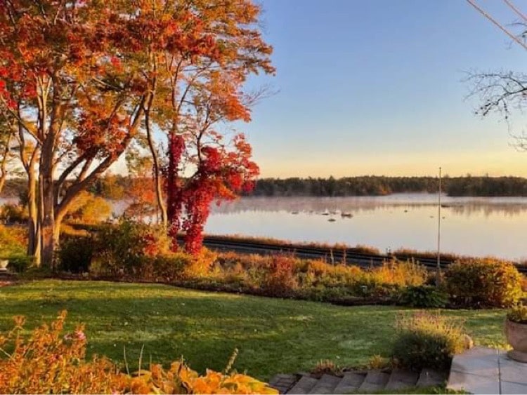 A serene view overlooking a calm lake surrounded by colorful autumn foliage. The area features lush green grass, seasonal trees with vibrant red and orange leaves, and tranquil waters reflecting the morning light.