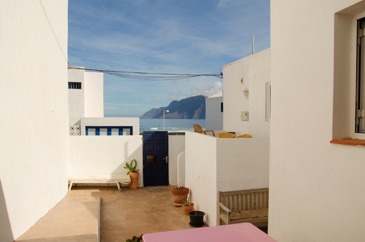 A bright outdoor terrace is visible, featuring seating and potted plants. The scene includes neighboring white structures and offers views of the ocean and Famara cliffs in the background under a clear blue sky.
