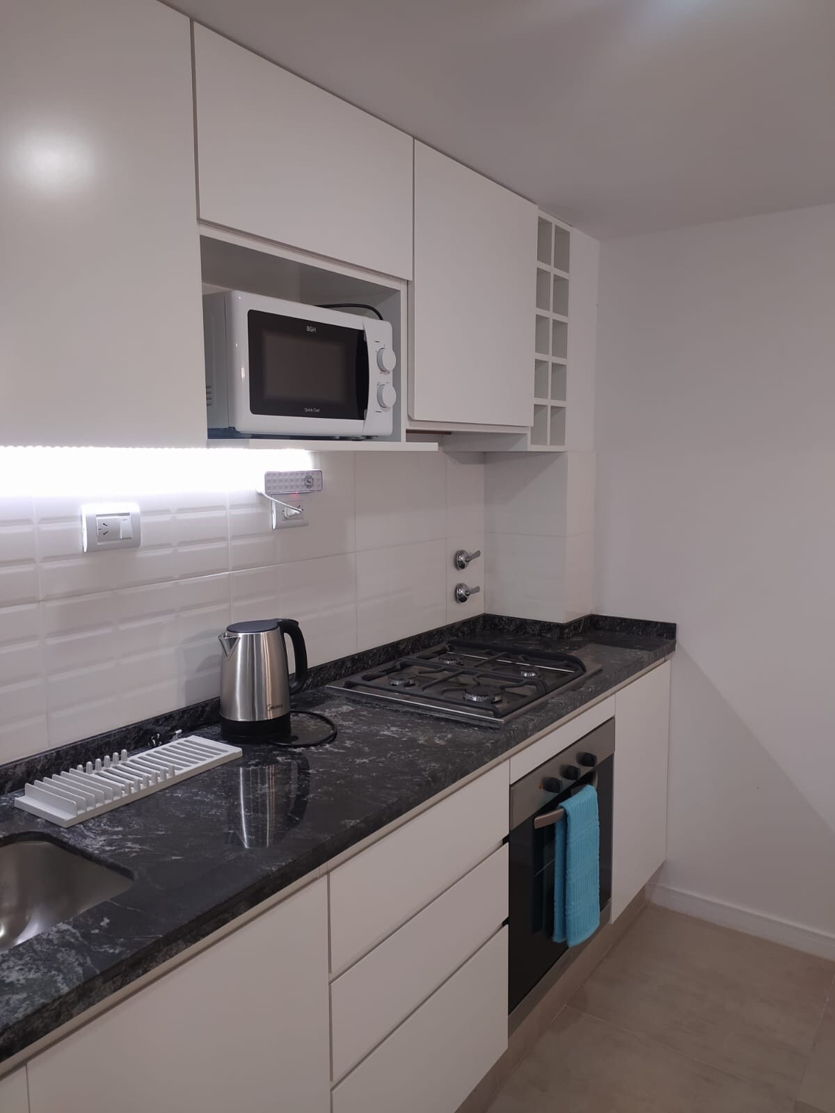 A modern kitchen features sleek white cabinetry and a dark granite countertop. Stainless steel appliances, including a microwave and kettle, are present. A cooktop is integrated into the countertop, and a small sink is located adjacent to it. Soft lighting highlights the cooking area.